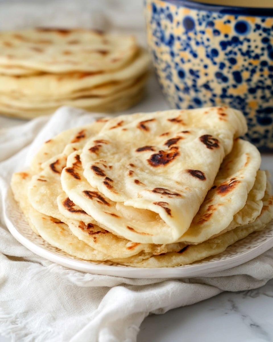 A stack of soft, round flatbreads is placed on a white plate with delicate patterns. The flatbreads are light beige with slightly charred dark brown spots scattered on the top surfaces. The stack shows around ten flatbreads, layered neatly one on top of the other with soft, slightly uneven edges, giving a fresh, homemade look. The background displays a white marbled texture with a blurred, pale yellow and blue bowl in the distance, and a white cloth nearby. Photo taken with an iphone --ar 4:5 --v 7