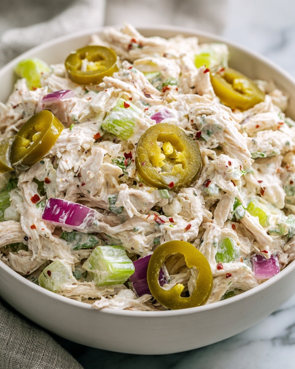 A close-up view of a white bowl filled with creamy chicken salad. The salad has shredded white chicken meat mixed with light green celery pieces and bits of purple-red onion. There are green pickled jalapeño slices on top, scattered over the creamy white dressing, which has small specks of herbs and red spices. The bowl sits on a white marbled surface with a soft rag beside it. photo taken with an iphone --ar 4:5 --v 7