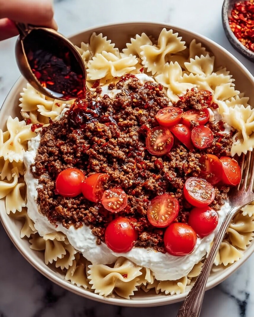 A white bowl filled with three clear layers, starting with pale yellow bowtie pasta at the bottom, topped with a thick layer of creamy white sauce, then a generous amount of cooked ground meat with a rich brown color and small bits of onion, finished with halved bright red cherry tomatoes sprinkled with dried green herbs on top. A silver fork rests inside the bowl, on the right side. The bowl is placed on a white marbled surface, with parts of another bowl and small dishes containing red and green spices visible around it. photo taken with an iphone --ar 4:5 --v 7