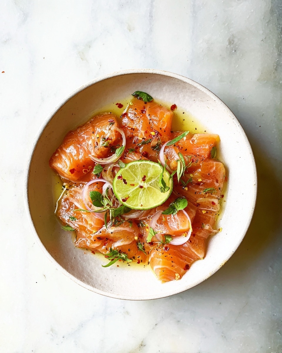 A round white bowl sits on a white marbled surface, filled with thin slices of raw salmon arranged loosely across the bowl’s base, each piece showing a smooth, shiny orange-pink texture. Scattered on top are small green leaves and thin pale rings of shallot or onion, adding touches of green and light purple. In the center rests a wedge of lime with bright green outer skin and translucent pale green flesh. The whole dish glistens with a light golden oil and is sprinkled with black pepper and red chili flakes, giving spots of black and red across the salmon. Photo taken with an iphone --ar 4:5 --v 7