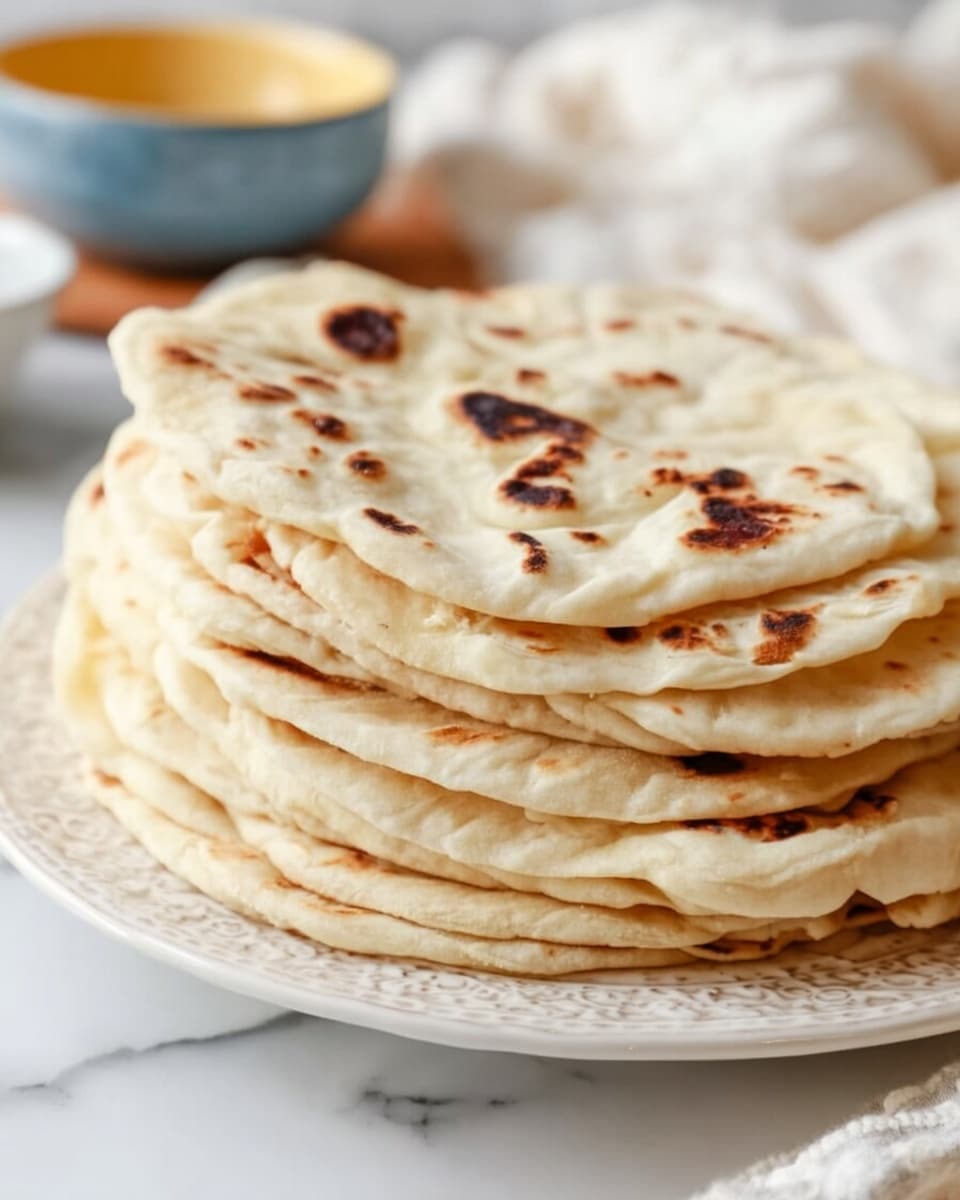 The image shows a stack of light golden flatbreads with dark brown char marks, placed on a white textured plate. The flatbreads are soft with a slightly puffed texture, three of them folded in half and stacked on top of four flat, open layers. The plate sits on a white marbled texture with a soft white cloth partially covering the flatbreads. In the background, there is a blurry stack of similar flatbreads and a portion of a large container with a blue and yellow speckled pattern. photo taken with an iphone --ar 4:5 --v 7
