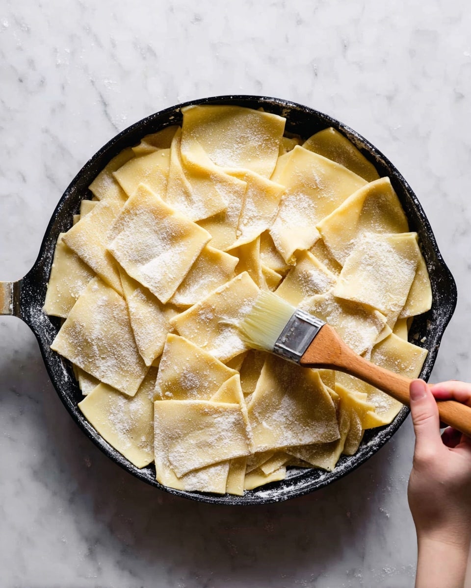 A round black pan filled with large, uneven square pieces of pasta dough layered slightly over each other, showing a pale yellow color with some areas dusted in white flour. A woman's hand is holding a wooden brush applying liquid on top of the pasta squares. The background is a white marbled texture. photo taken with an iphone --ar 4:5 --v 7
